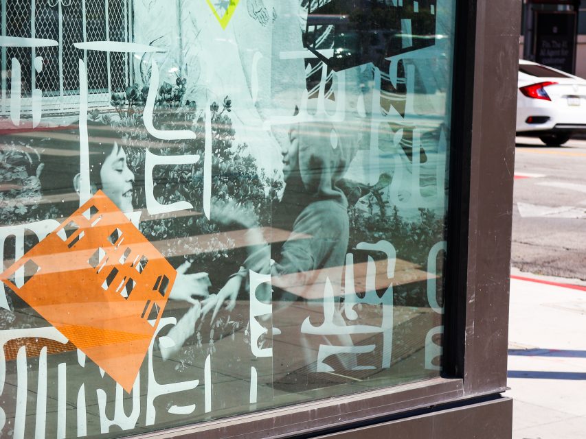 A bus stop shelter with geometric and abstract white patterns on glass, reflecting two people interacting behind the glass near a city street.