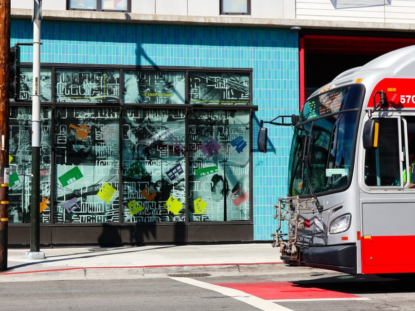 A city bus is stopped at a street corner near a building with turquoise tiles and abstract art on the windows.