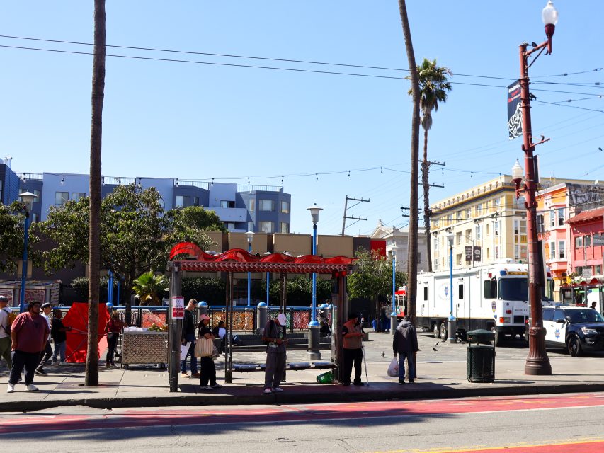 People gather at a bus stop with a red roof in an urban area; buildings, trees, and utility vehicles are visible in the background.