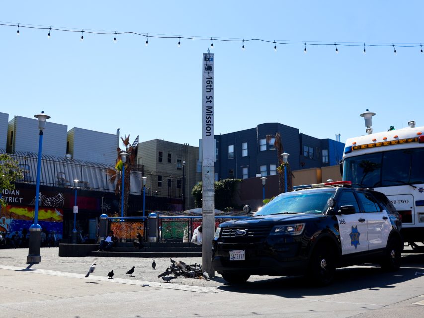 A police car is parked by a pole labeled "16th St Mission" near a bus, with pigeons on the pavement and colorful buildings in the background.