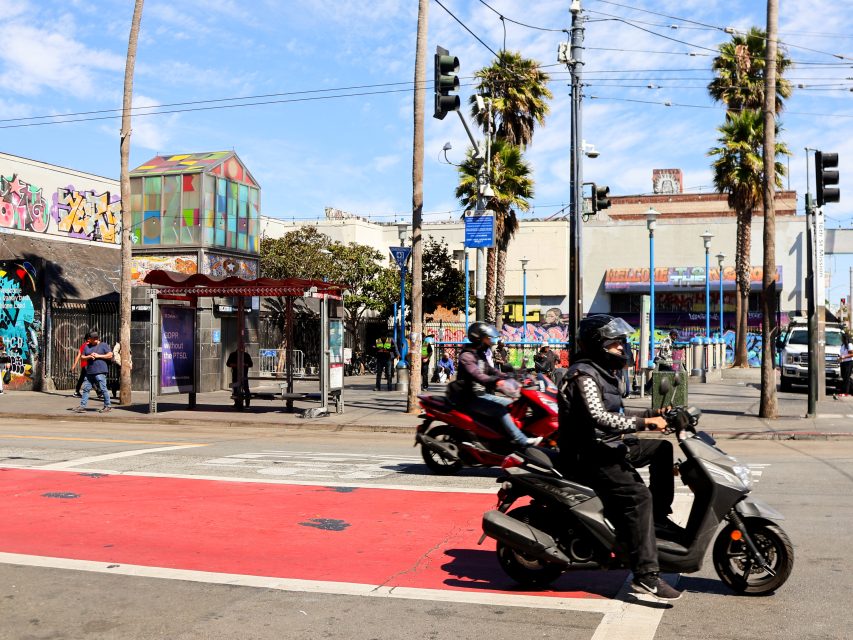 Two people on scooters ride through a crosswalk at a city intersection with graffiti-covered buildings, palm trees, and a bus stop in the background.