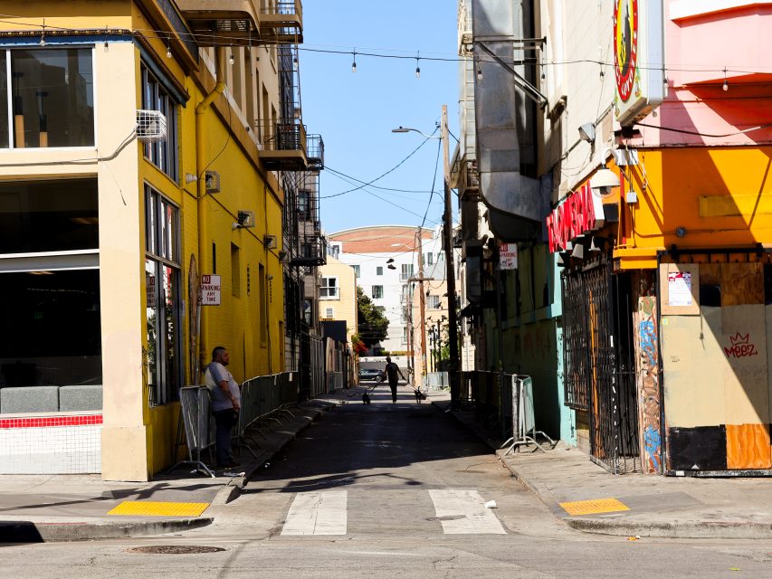 A narrow urban alleyway between yellow and orange buildings with a few people standing and walking under clear daylight.