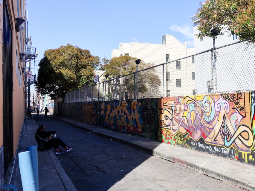Two people sit on the ground in a sunlit alley lined with colorful graffiti-covered walls and a chain-link fence, with buildings and trees in the background.