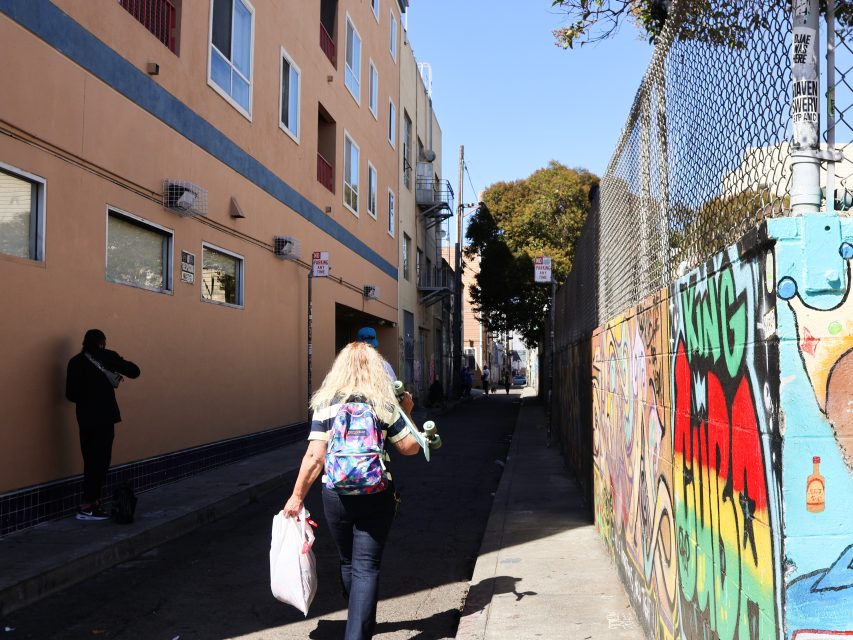 A person with a backpack and skateboard walks down a narrow alleyway lined with colorful graffiti and apartment buildings, as another person stands near the wall.