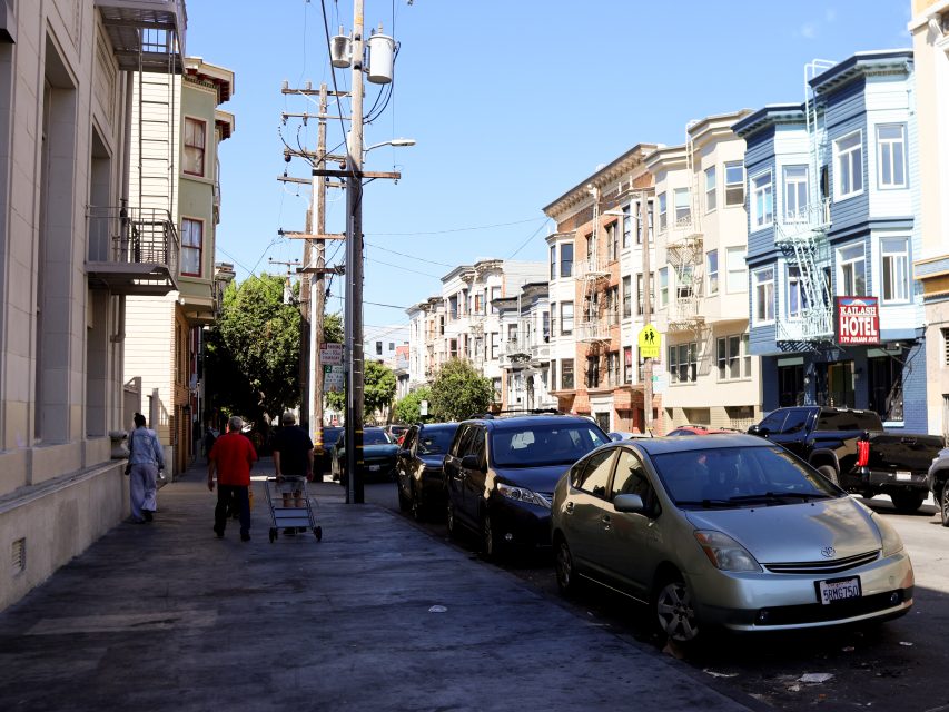 A city street with parked cars, utility poles, and multi-story apartment buildings; two people walk on the sidewalk, one pushing a cart.