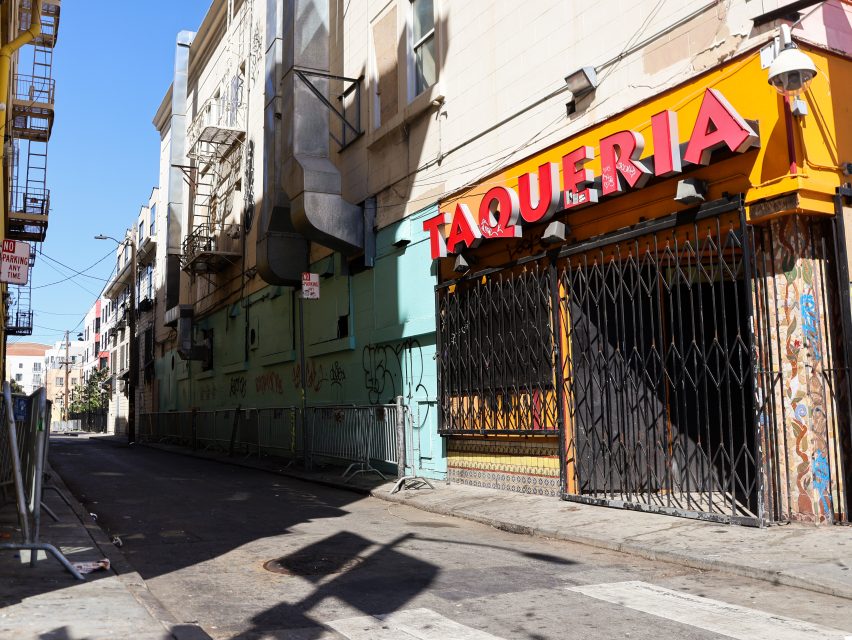 A closed taqueria with a yellow and red sign on a narrow, empty city street. Metal gates cover the entrance and graffiti marks the walls.