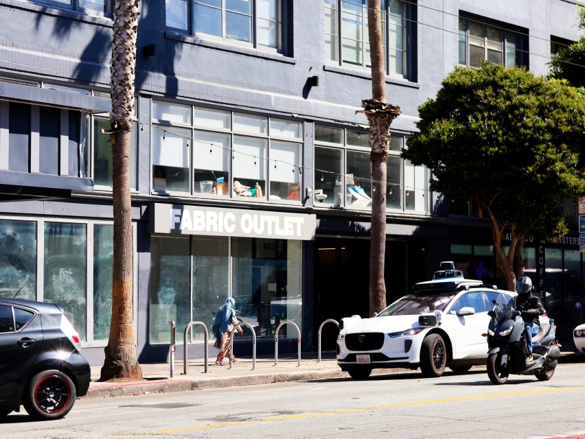 A person walks a bike in front of a "Fabric Outlet" store on a sunny street with parked cars, motorcycles, and palm trees.