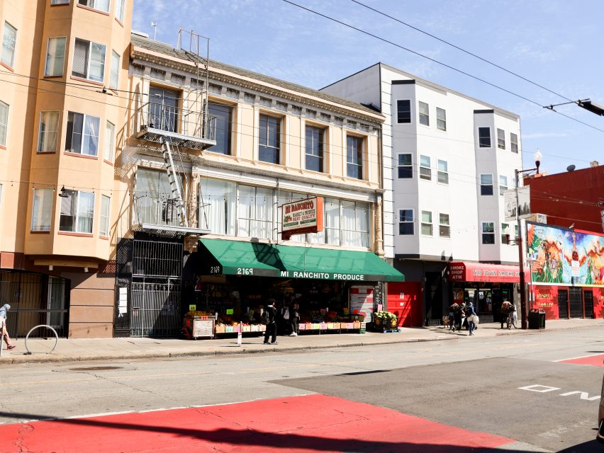 A street view of a produce market called "Mi Ranchito Produce" with a green awning, located on the ground floor of a multi-story building. People are walking on the sidewalk.
