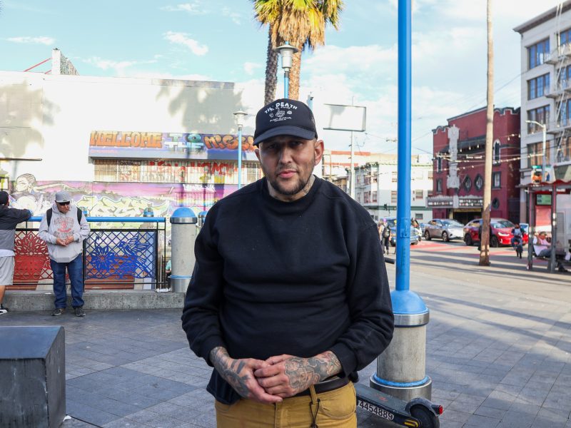 A man with tattoos on his hands stands in an urban plaza, wearing a black sweatshirt, tan pants, and a baseball cap. Buildings, a palm tree, and people are in the background.