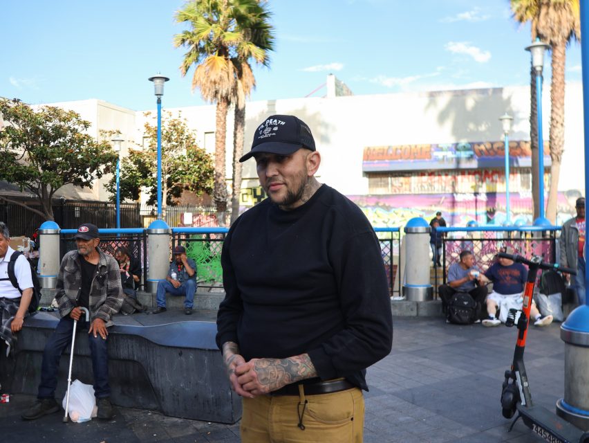 A man in a black cap and sweater stands in an urban park area, with people sitting nearby and graffiti on a wall in the background.
