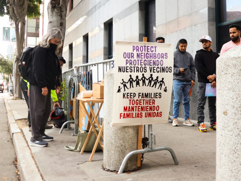 People stand in line on a city sidewalk near a sign that reads "Protect Our Neighbors, Keep Families Together" in English and Spanish.