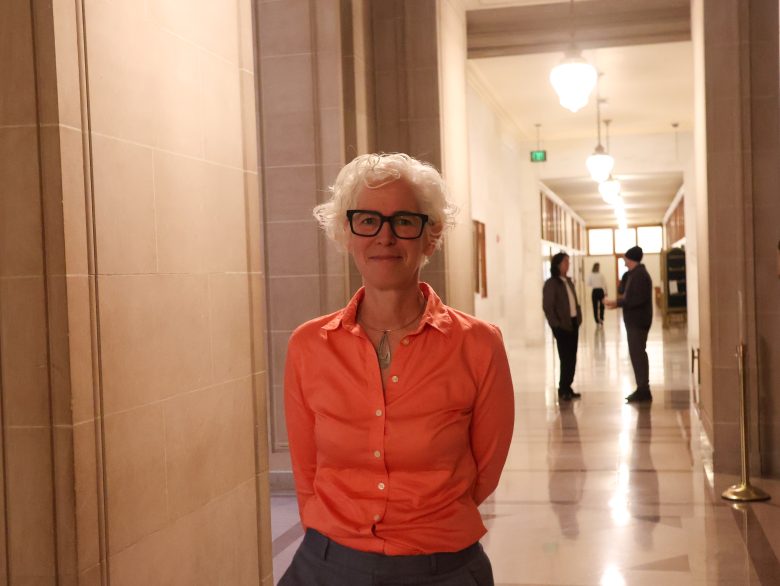 A person with short white hair and glasses stands in a hallway with cream-colored walls and bright overhead lights; several people are seen in the background.