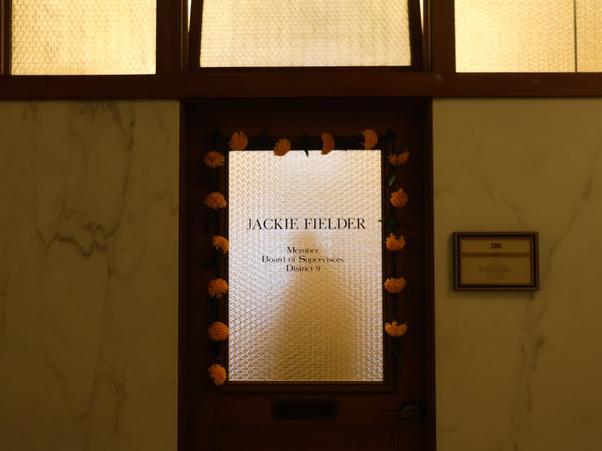 A wooden door with a frosted glass window reads "Jackie Fielder, Member, Board of Supervisors, District 9," decorated with orange flowers; a plaque beside it reads "280 Reception.