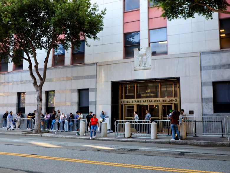People stand in a line outside the United States Appraisers Building, with metal barriers and trees along the sidewalk.