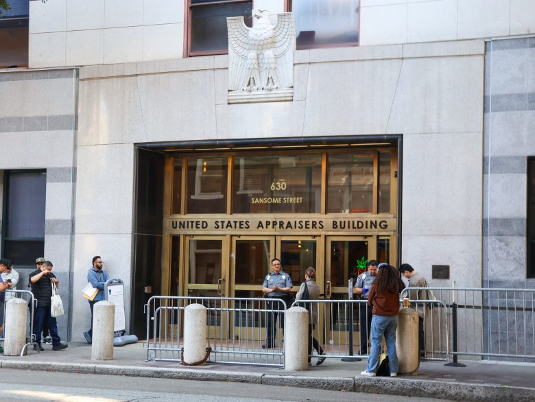 People wait outside the entrance of the United States Appraisers Building at 630 Sansome Street, where ICE security personnel and metal barricades are present.