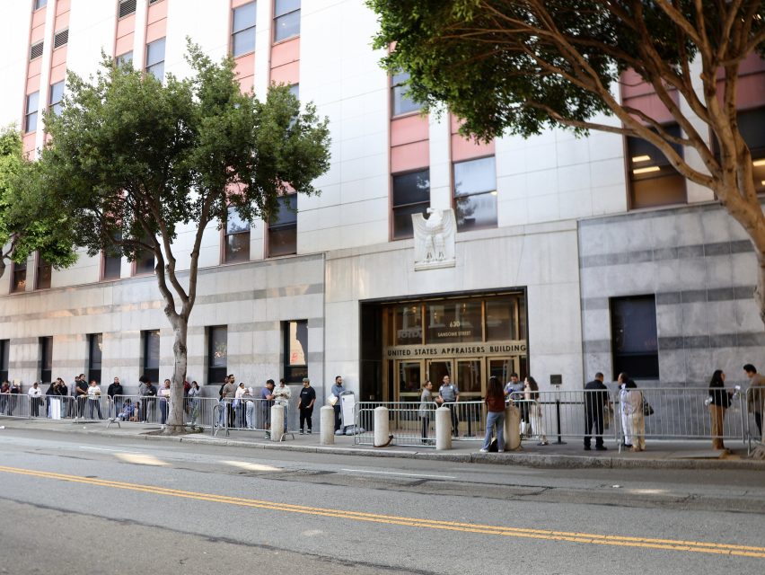People stand in a line outside the United States Appraisers Building on a city street, with trees and metal barricades along the sidewalk.