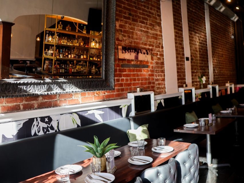 A restaurant with exposed brick walls, a large mirror reflecting a bar, set tables with plates and glasses, and cushioned seating along the wall.
