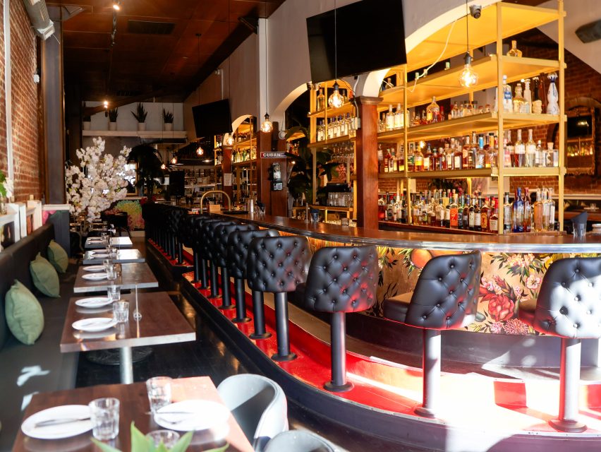 A brightly lit restaurant interior with a long bar, black tufted bar stools, set dining tables, and shelves filled with bottles behind the bar.