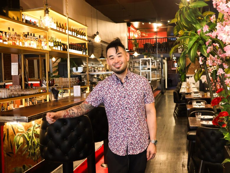 A man stands and smiles inside a Caribbean-inspired restaurant with a bar on one side, tables set for dining, and lush plants decorating the space.