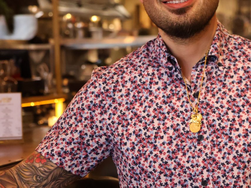 A person wearing a floral button-up shirt and gold necklaces, with a tattooed arm, standing indoors in a warmly lit setting.