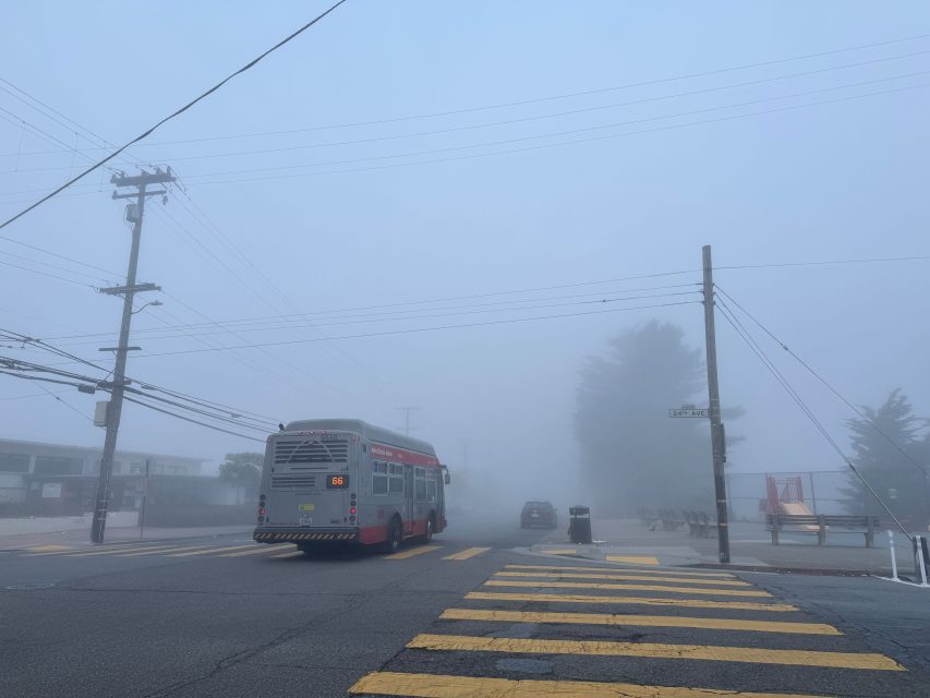 A city bus drives down a foggy street with yellow crosswalk stripes, power lines overhead, and a few parked cars and benches barely visible through the mist, evoking a sense of quiet recall.