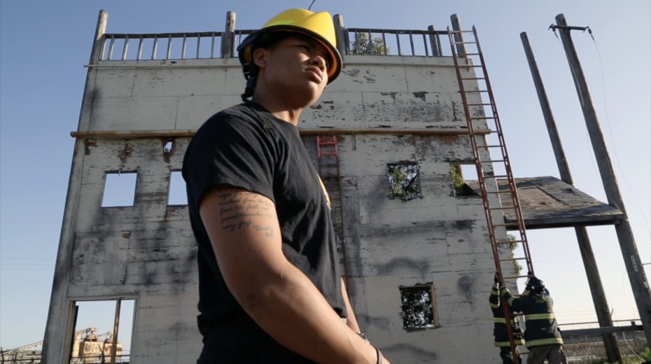 Person wearing a yellow hard hat and black shirt stands in front of a weathered building with ladders and firefighters visible in the background.