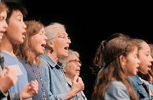 A diverse group of children and older adults, all wearing blue tops, stand in a row singing together on stage.