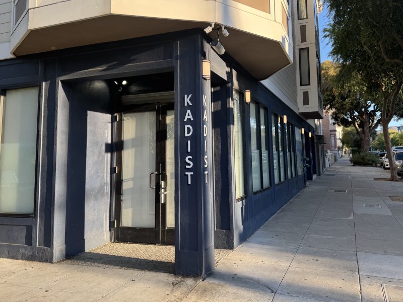 A dark blue building corner with large windows and a glass door displays the word "KADIST" in white vertical letters; the sidewalk and trees line the adjacent street.