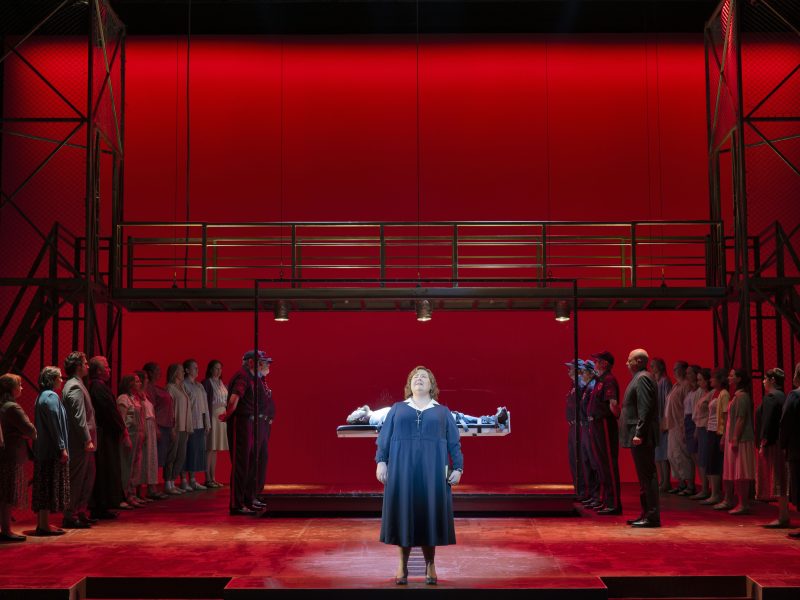 A woman stands center stage in front of a lit platform with a person lying down, surrounded by uniformed guards and two groups of onlookers, set against a vivid red backdrop.