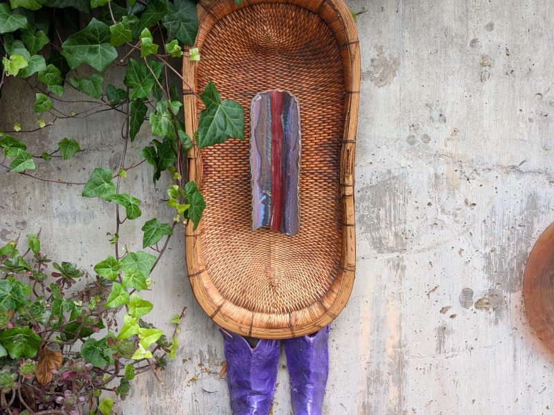 A woven basket with purple boots and a painted wooden plank attached is mounted vertically on a concrete wall, surrounded by green ivy.