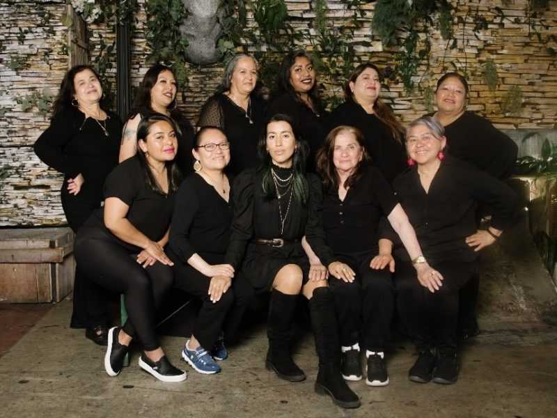 A group of eleven women dressed in black pose together for a group photo in front of a stone wall decorated with greenery.