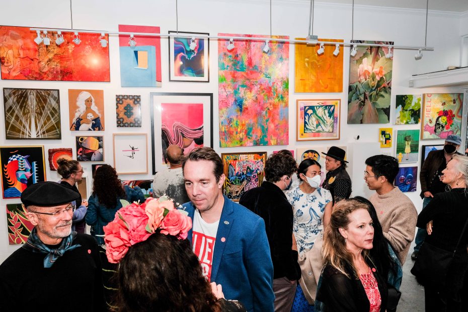 A group of people view and discuss colorful artwork displayed on the walls of a gallery during an art exhibition.