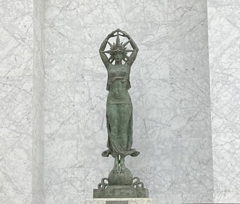 Bronze statue of a woman standing with arms raised, holding a star-shaped object, set against a marble wall background.