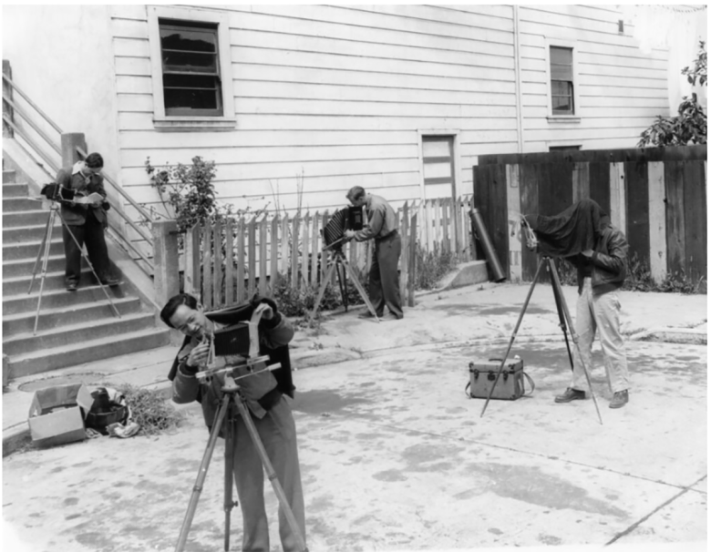 Four people operate vintage cameras on tripods in an outdoor urban setting, reminiscent of scenes often found in museums, with one person using a camera on steps and another beneath a dark cloth.