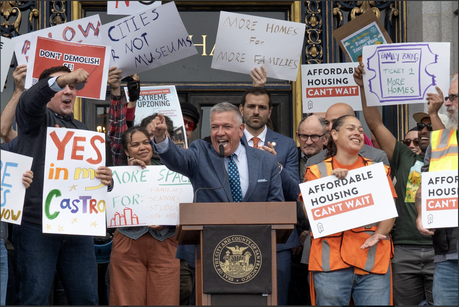 A man speaks at a podium while a diverse group behind him holds signs advocating for affordable housing, more homes in the city, and support for the upzoning plan.