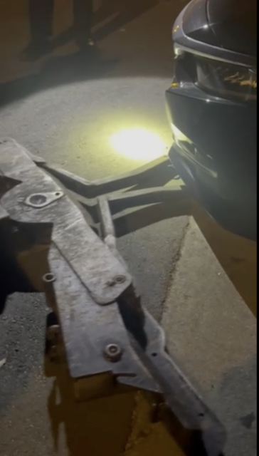 A car in front of a tow truck's lift at night, illuminated by a bright flashlight on an asphalt surface.