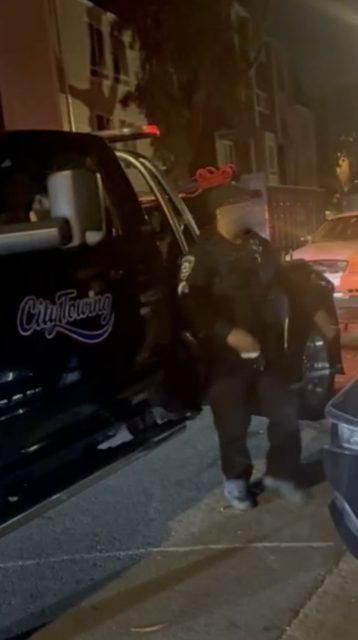 A police officer stands next to a tow truck labeled "City Towing" at night near parked vehicles on a city street.