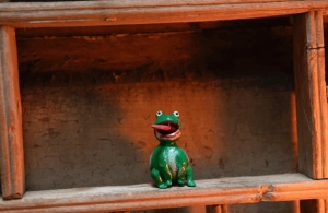A small green frog figurine with an open mouth sits on a wooden shelf against a rustic background.