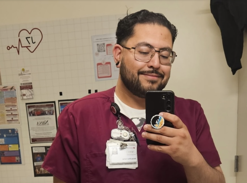 A man wearing maroon scrubs and badges takes a selfie in front of a mirror in what appears to be a medical or hospital setting.