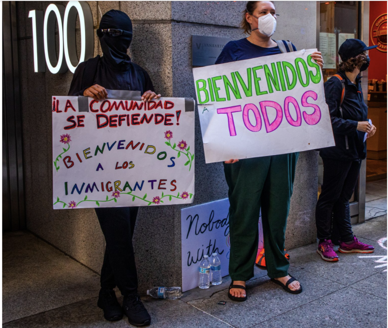 Two people stand outside holding signs in Spanish welcoming immigrants. One wears a mask and holds a sign that says "¡Bienvenidos a los inmigrantes!" and the other holds "Bienvenidos a todos.