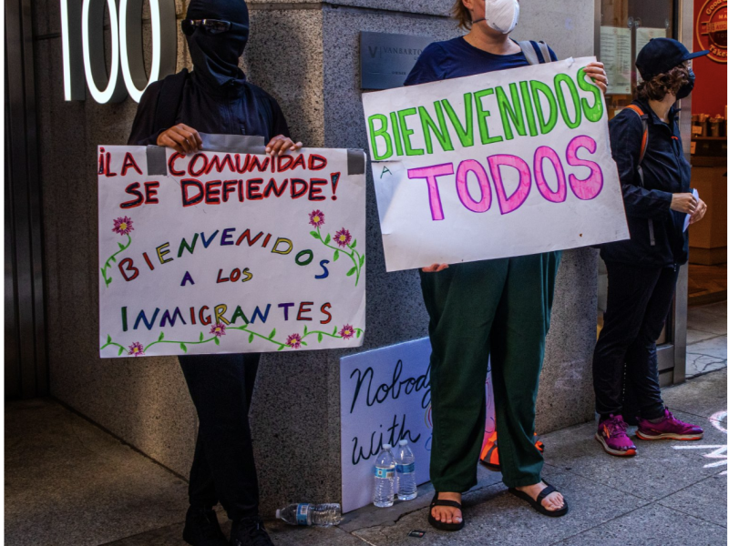 Two people stand outside holding signs in Spanish welcoming immigrants. One wears a mask and holds a sign that says "¡Bienvenidos a los inmigrantes!" and the other holds "Bienvenidos a todos.