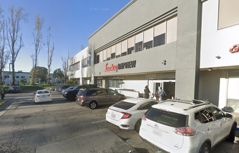 Parking lot in front of a Lucky Bayview grocery store, with several parked cars and people entering or exiting the store on a sunny day.