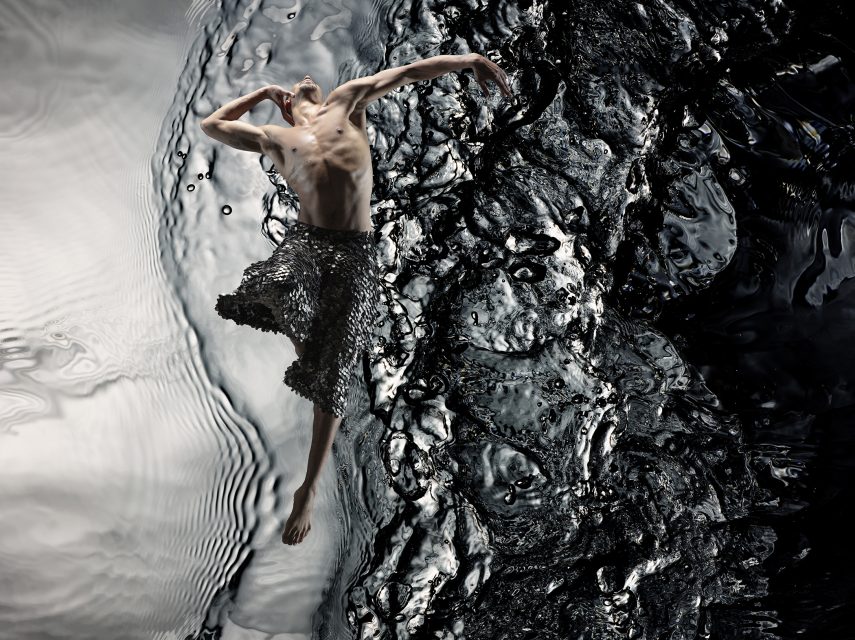 A shirtless person in a metallic skirt leaps gracefully against a swirling black and silver water background.