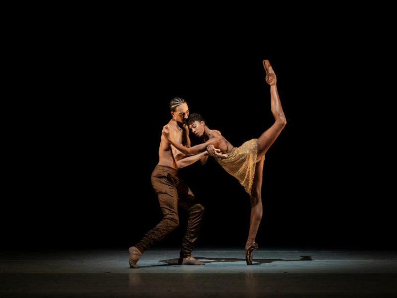 Two ballet dancers perform on stage; one balances en pointe with her leg extended high, while the other supports her. Both wear neutral-toned costumes against a dark background.