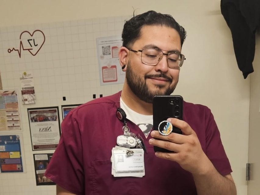 A person wearing maroon scrubs and a name badge takes a selfie in a room with a whiteboard and various notices on the wall, highlighting their professional environment and protected status.