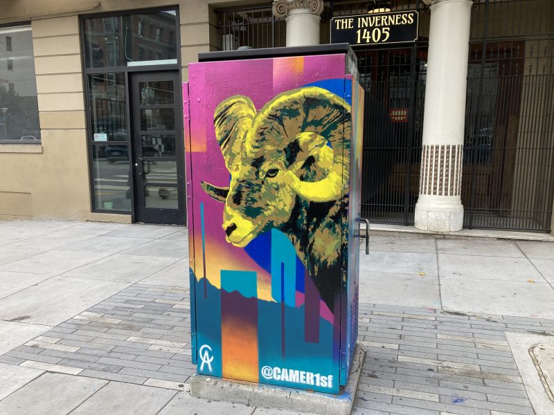 A painted utility box on a city sidewalk features a stylized ram’s head with a colorful, abstract urban background and the text "@CAMER1sf.