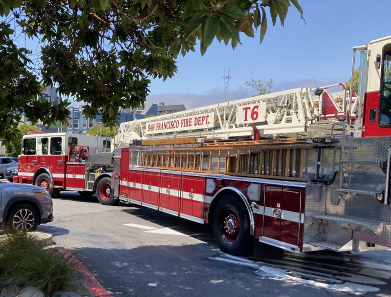 Two red and white fire trucks from the San Francisco Fire Department, one with a large ladder labeled T6, are parked on a city street near trees and buildings.