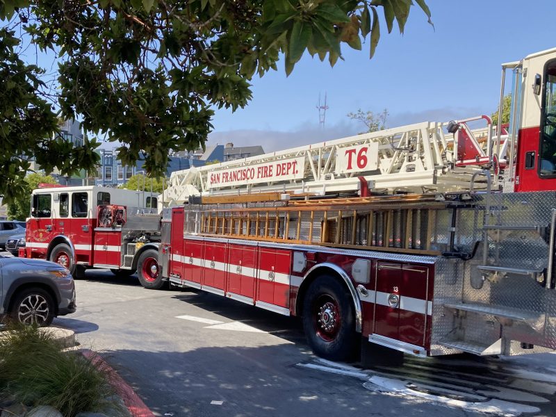 Two red and white fire trucks from the San Francisco Fire Department, one with a large ladder labeled T6, are parked on a city street near trees and buildings.