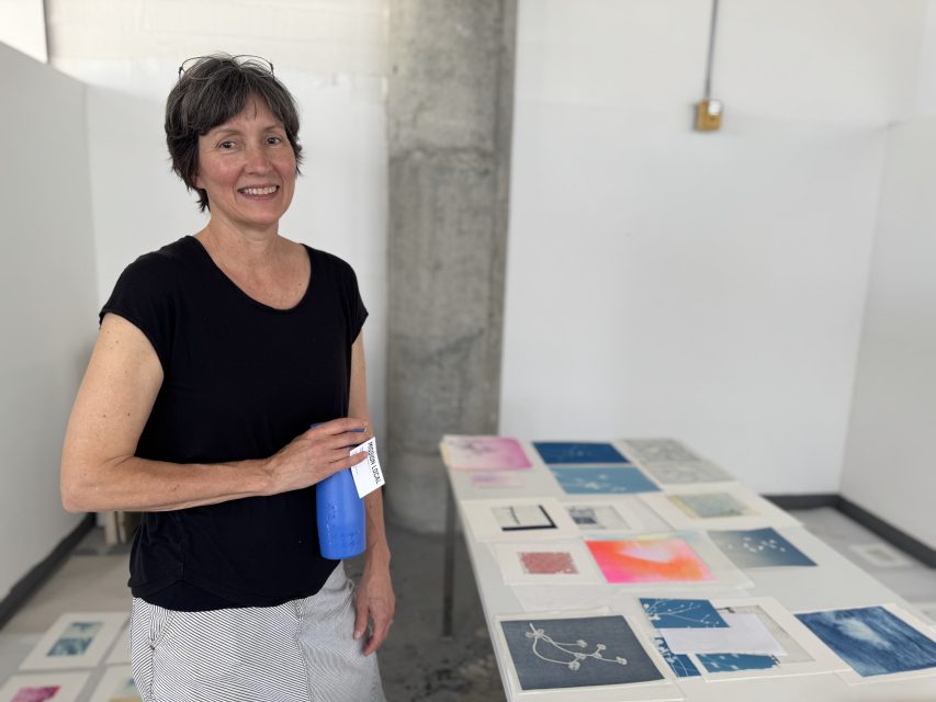 A woman stands indoors next to a table displaying various abstract art pieces, holding a blue water bottle and smiling at the camera.