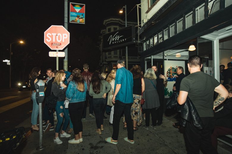 A group of people gather outside an art gallery at night near a stop sign on a city sidewalk.